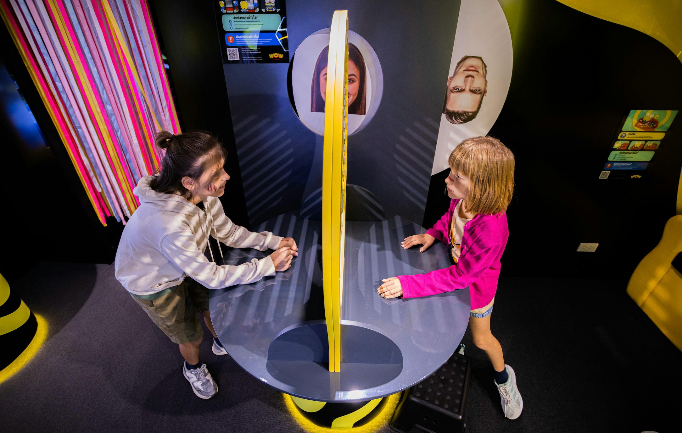 A child interacts with an exhibit featuring a yellow divider and images of faces, inside a brightly lit interior.