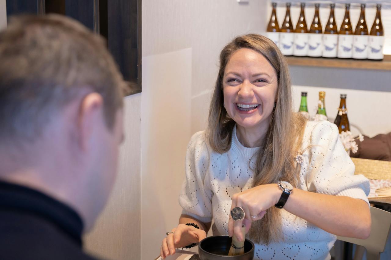 A woman smiling while stirring a bowl, with bottles displayed on shelves in the background.