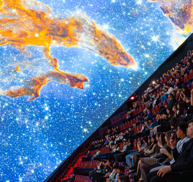 사람들은 천체투영관에 앉아 커다랗고 구부러진 화면에서 별과 성운의 생생한 우주 전시를 보고 있습니다.