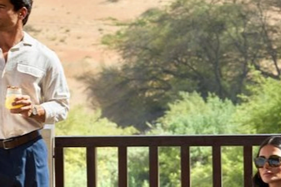 A man and a woman relaxing on a shaded terrace with seating, overlooking a desert landscape with scattered trees.