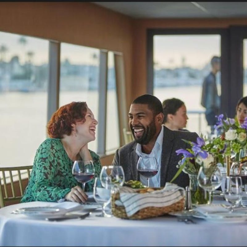 A man and a woman smiling at each other while seated at a dining table with wine glasses, flowers, and a bread basket. Other people in the background.