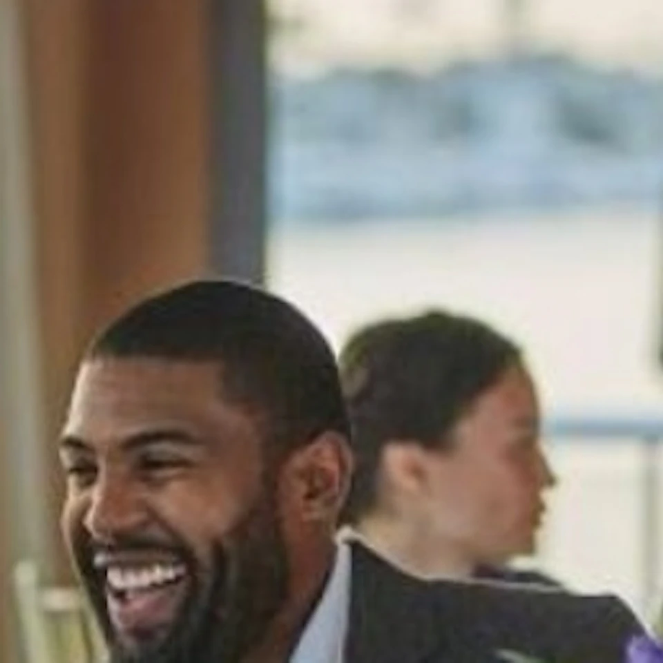 A man and a woman smiling at each other while seated at a dining table with wine glasses, flowers, and a bread basket. Other people in the background.