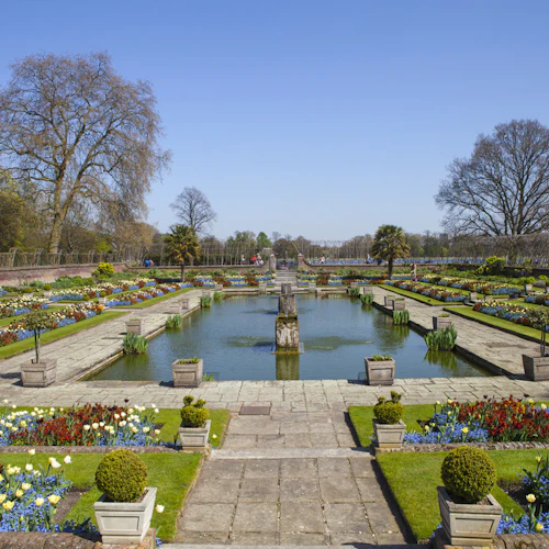 Formell trädgård med symmetriska rabatter och en central damm, omgiven av gångar, träd och välskötta buskar.