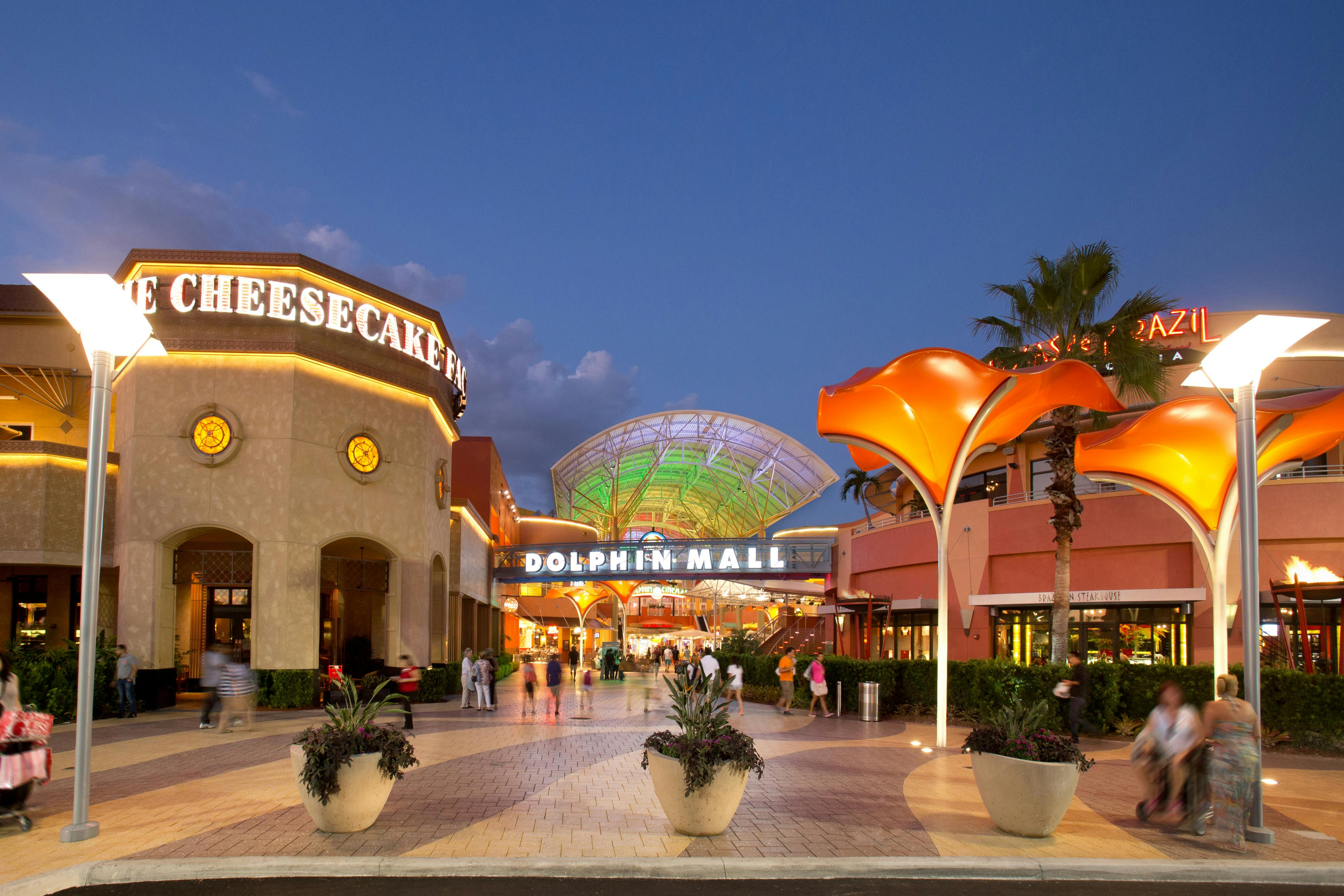 Outdoor evening view of Dolphin Mall entrance with illuminated signage, vibrant lighting, and visitors walking nearby.