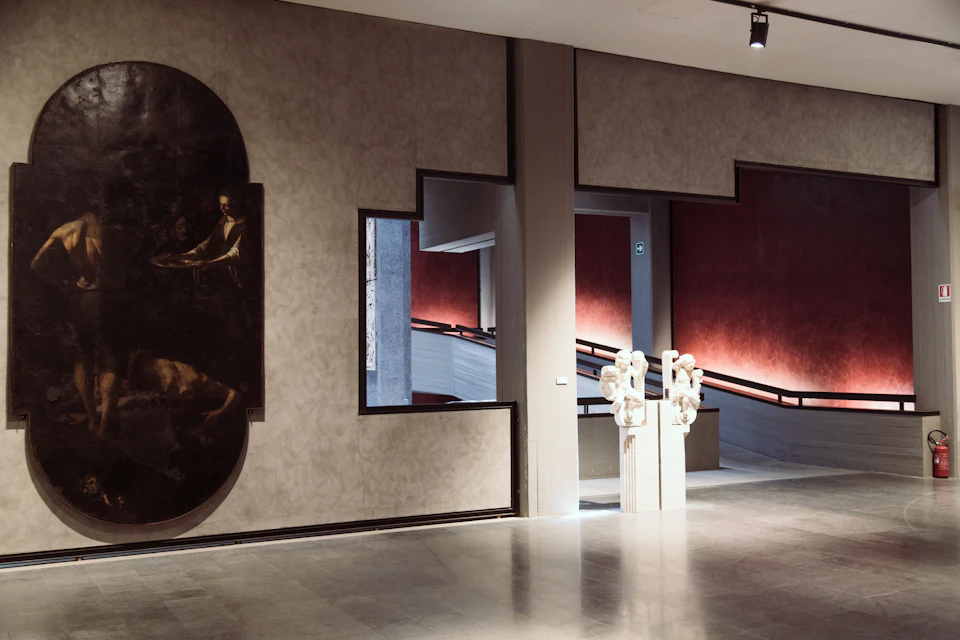 Modern museum interior with abstract wall design, a large painting on the left, and a sculpted statue near a staircase on the right.