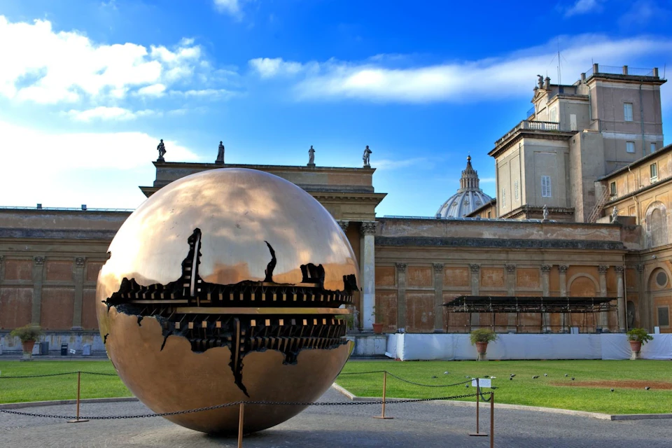 A large bronze sculpture of a sphere with internal structure is displayed outdoors near classical architecture on a sunny day.