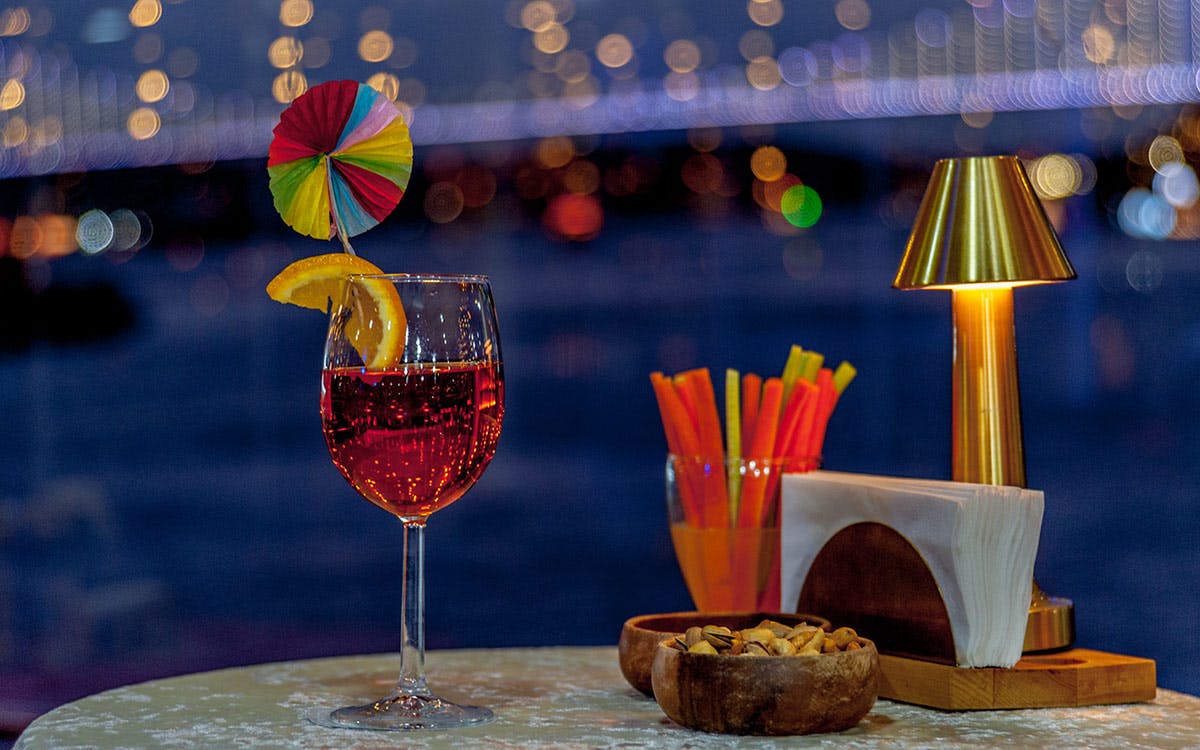 A wine glass with a red drink, orange slice, and colorful umbrella, nuts in a bowl, straws, and tissue on a table. Blurred lights in the background.