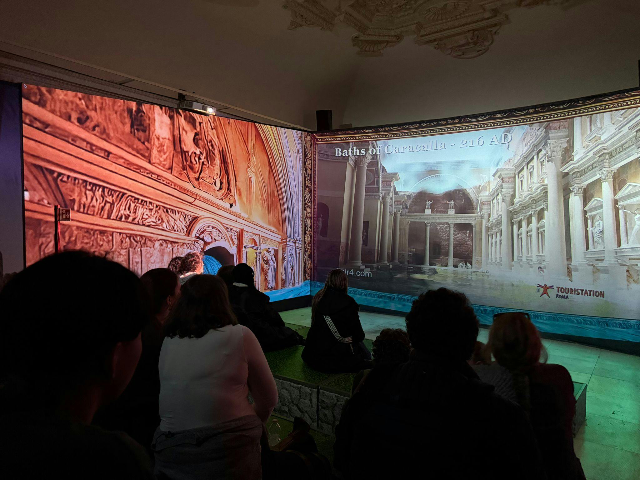 Un gruppo di persone sedute in una stanza buia, guarda una proiezione di un'antica architettura romana intitolata "Terme di Caracalla - 216 d.C.".