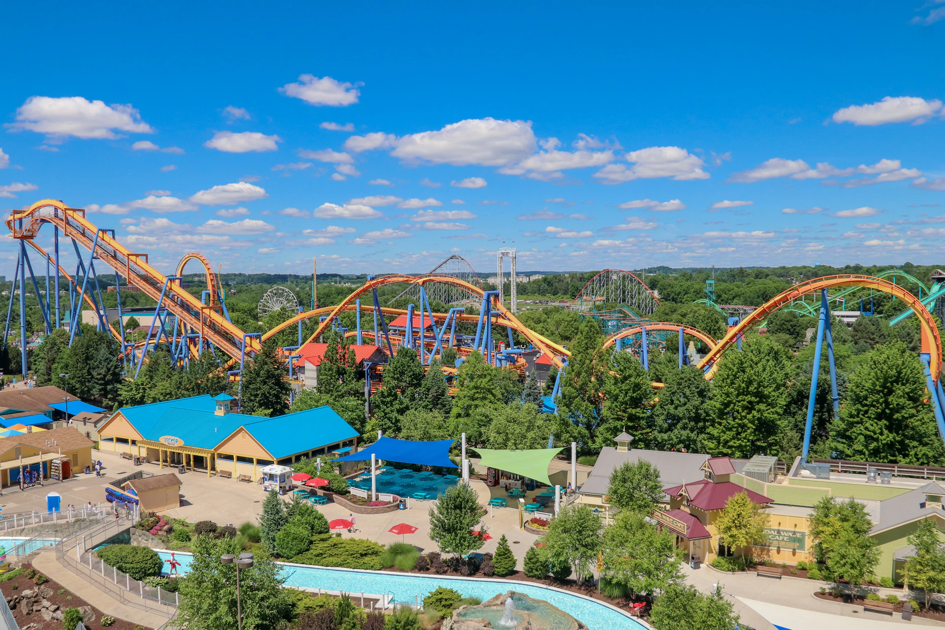 A theme park with colorful roller coasters, a water feature, and surrounding trees under a blue sky with scattered clouds.