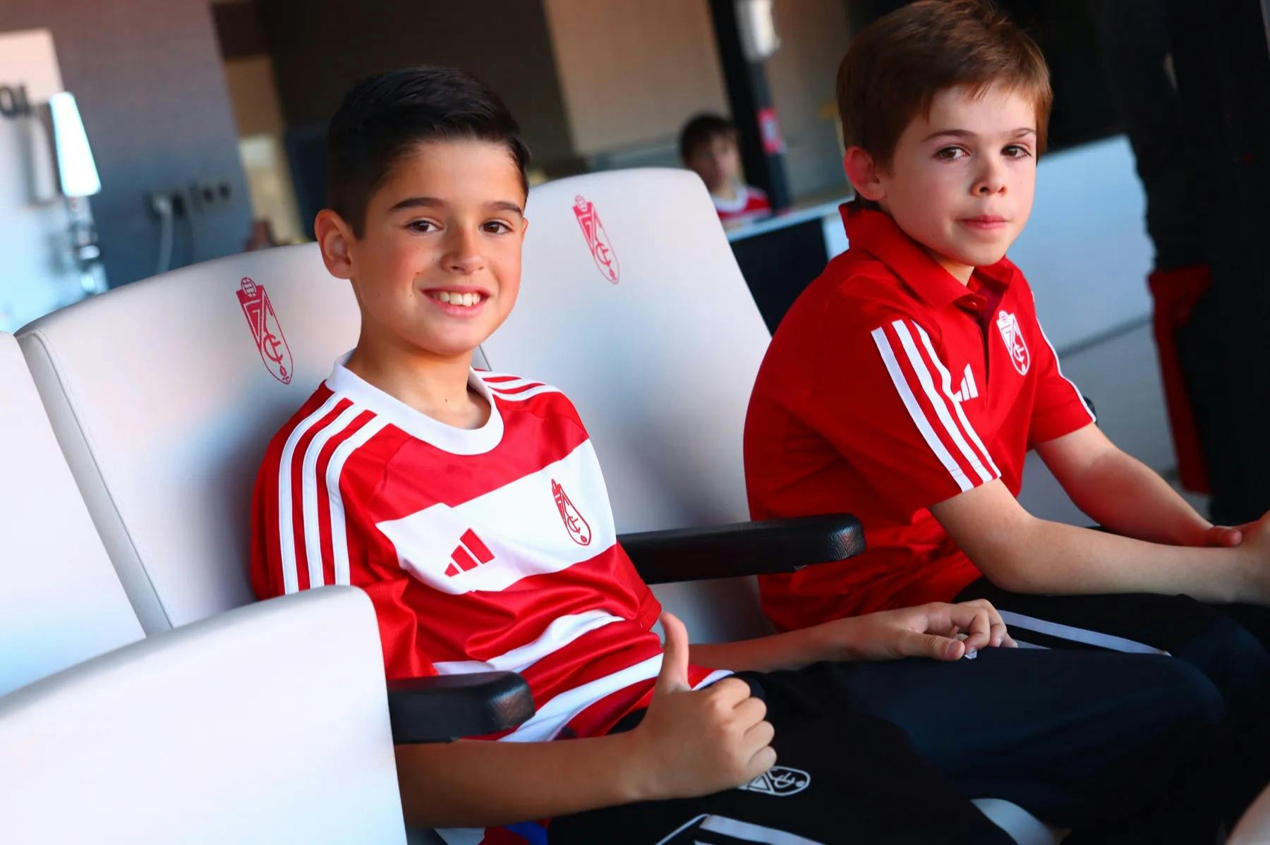 빨간색과 흰색 스포츠 유니폼을 입은 두 명의 소년이 흰색 좌석에 앉아 한 명은 엄지손가락을 치켜들고 미소를 짓고, 다른 한 명은 앞을 내다보고 있습니다.