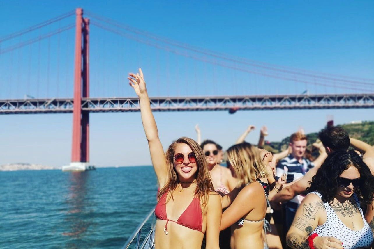 A young woman and a group of young people having fun on a boat party in Lisbon, with Ponte 25 de Abril in the background.