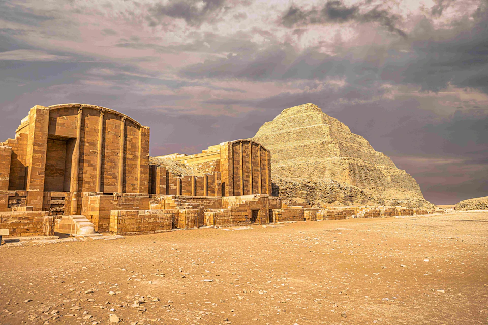A stepped pyramid and ancient stone structures under a cloudy sky in a desert landscape.