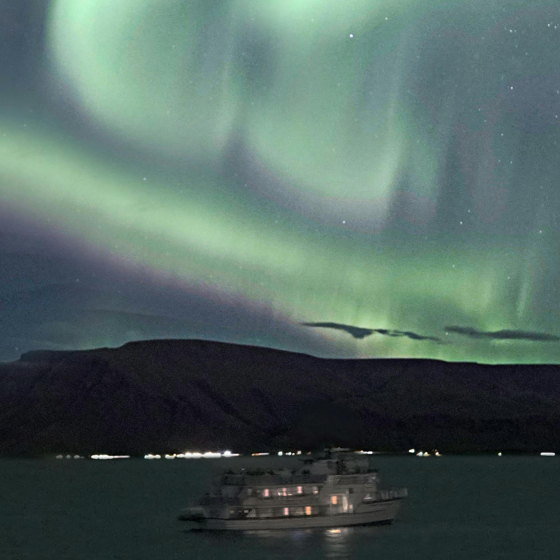 A boat on calm water under a vivid display of green and pink Northern Lights, with hills in the background against a night sky.