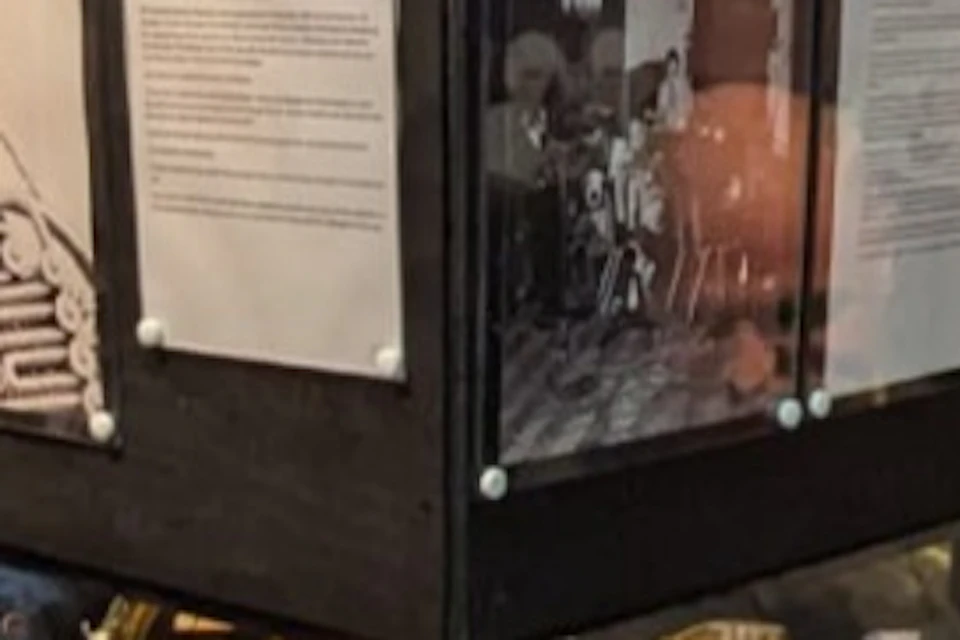 A group of people observing a glass display case containing a musical instrument, with some taking photos on their phones.
