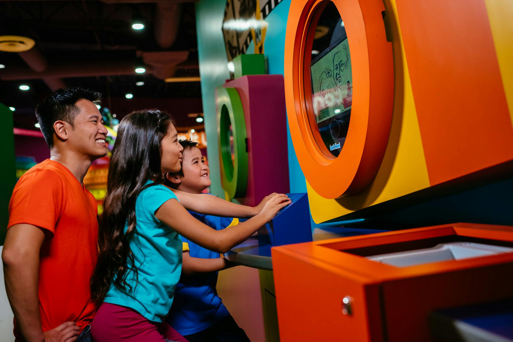 A man and two children interact with colorful arcade games, smiling and focused on the screens in a brightly lit indoor setting.