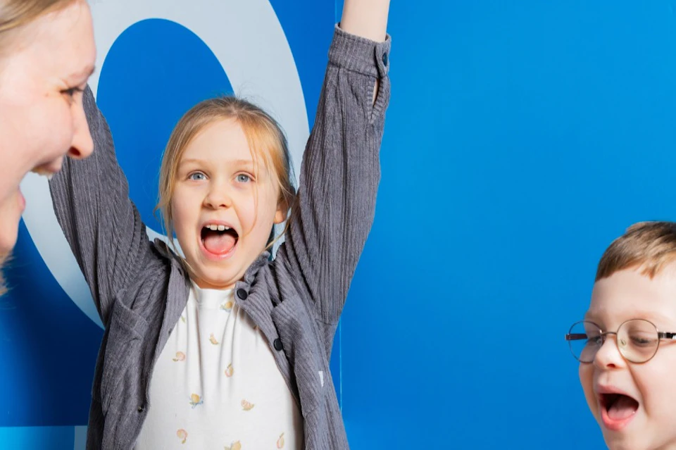 A woman and two children enthusiastically play a game on a table with colorful circles, against a blue background.