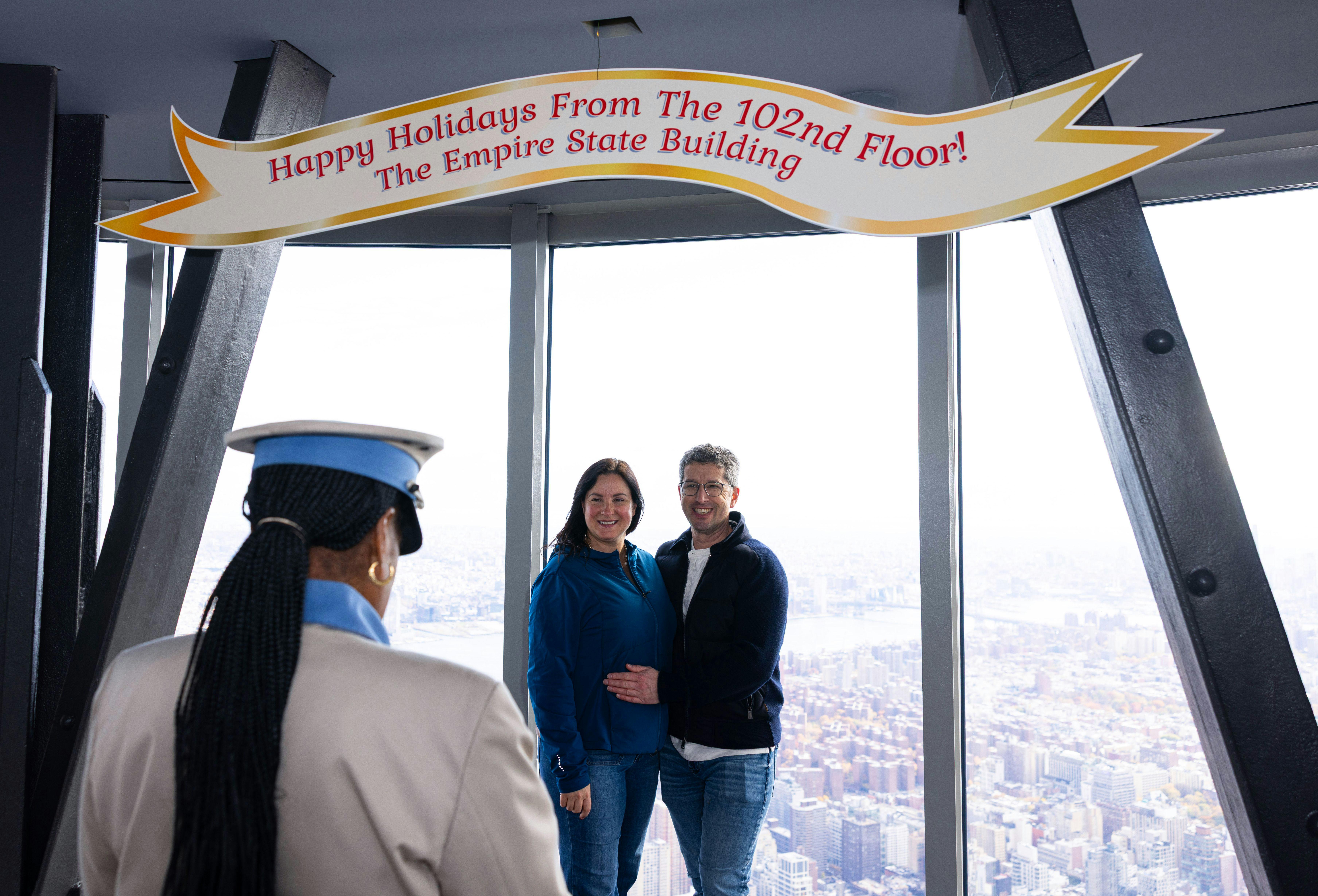Δύο άτομα ποζάρουν για μια φωτογραφία κάτω από ένα πανό στον 102ο όροφο του Empire State Building, με θέα στην πόλη από πίσω.