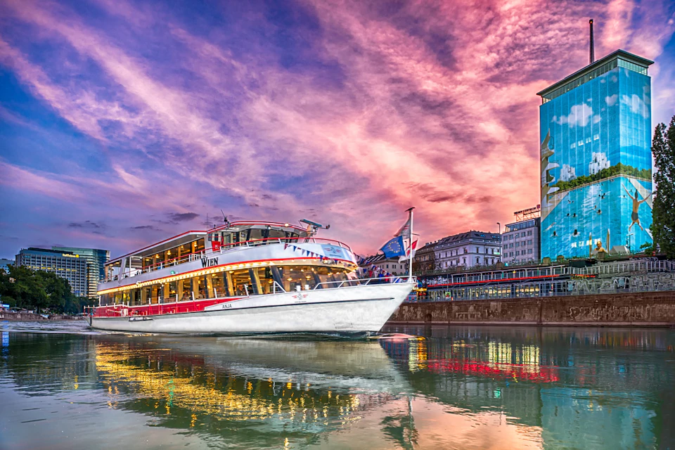 A white tour boat with "Wien" written on it travels through water at sunset, with buildings and reflections visible onshore.