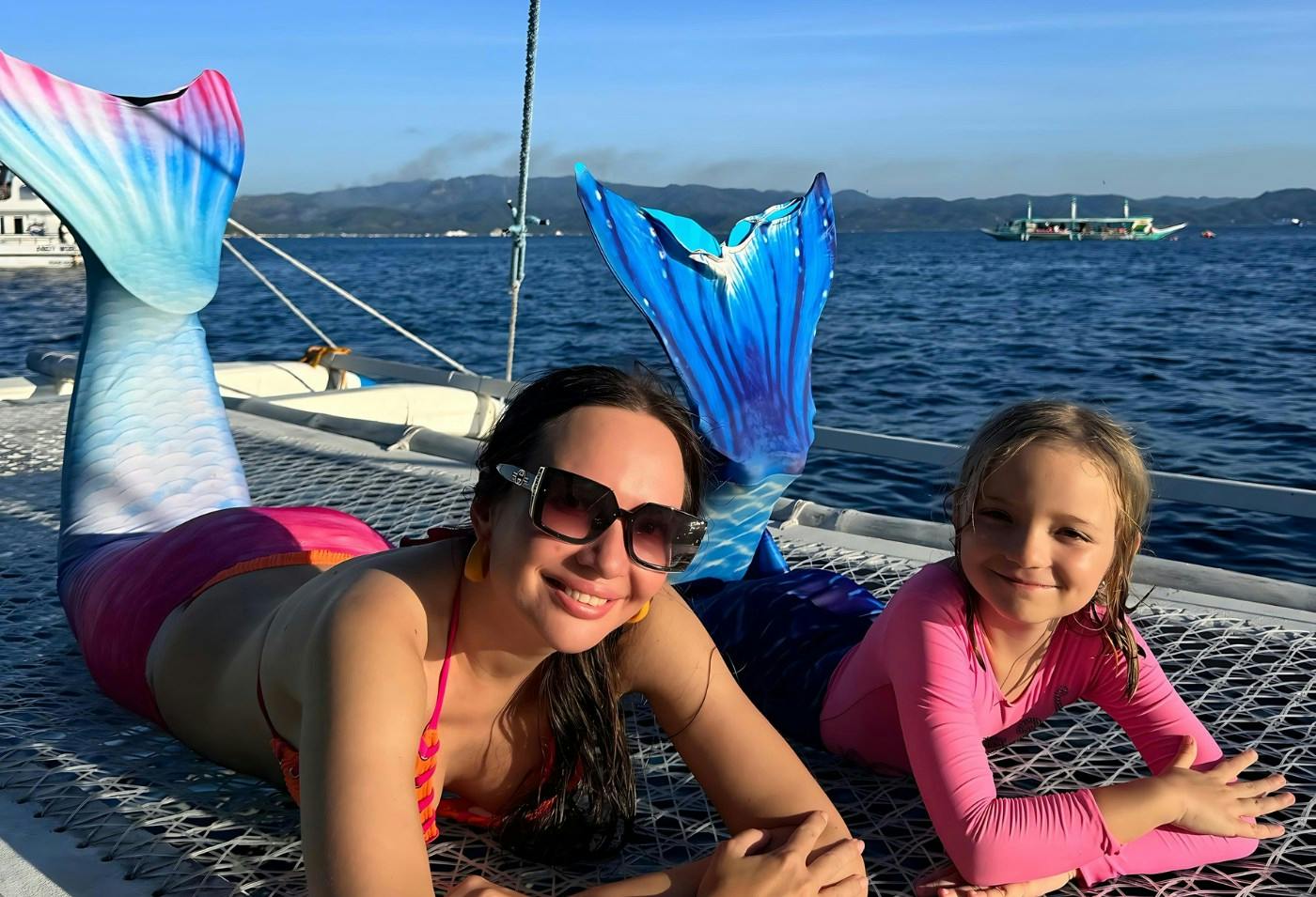 Two people enjoying a sunny day on a boat, one wearing a mermaid tail costume. A scenic ocean view is in the background.
