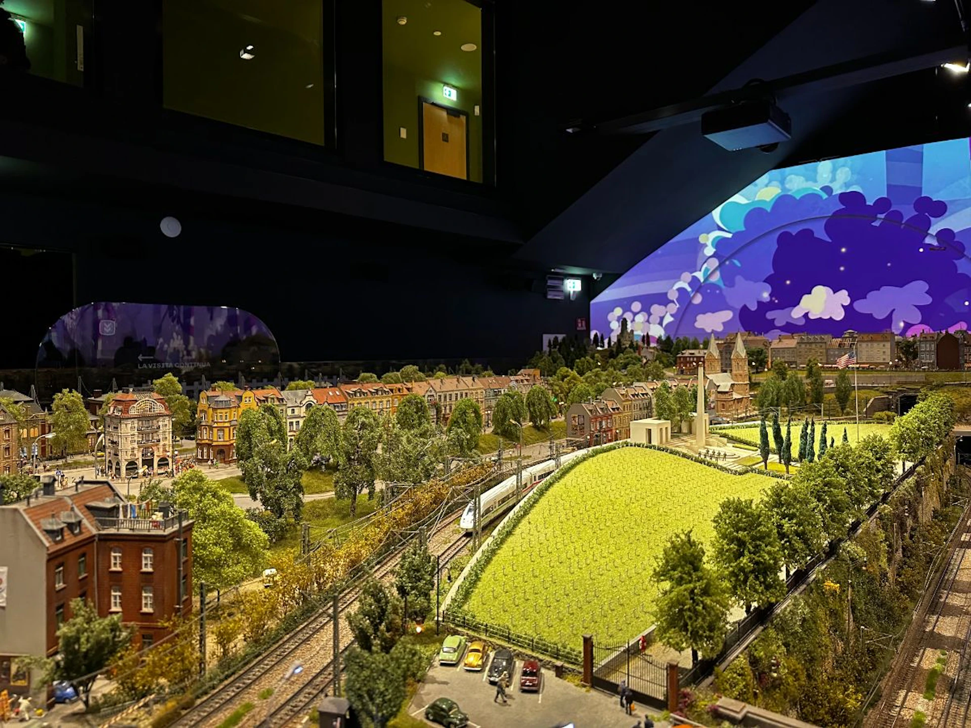Intricate miniature model of a town with detailed buildings, greenery, a train track, and a large backdrop painting of clouds.