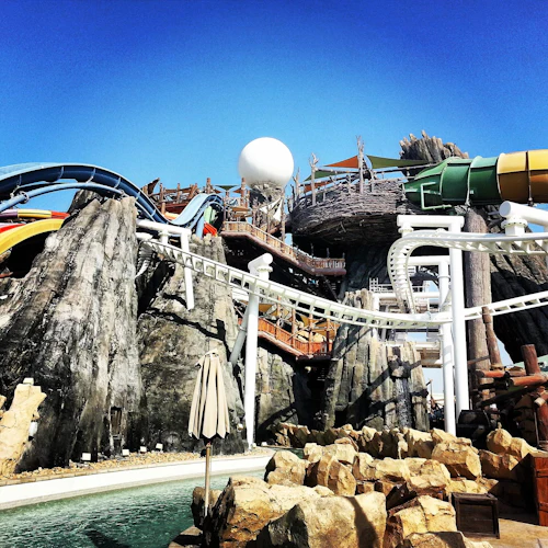 Vergnügungsparkszene mit Wasserrutschen, Felsstrukturen und Achterbahnschienen vor einem klaren blauen Himmel.