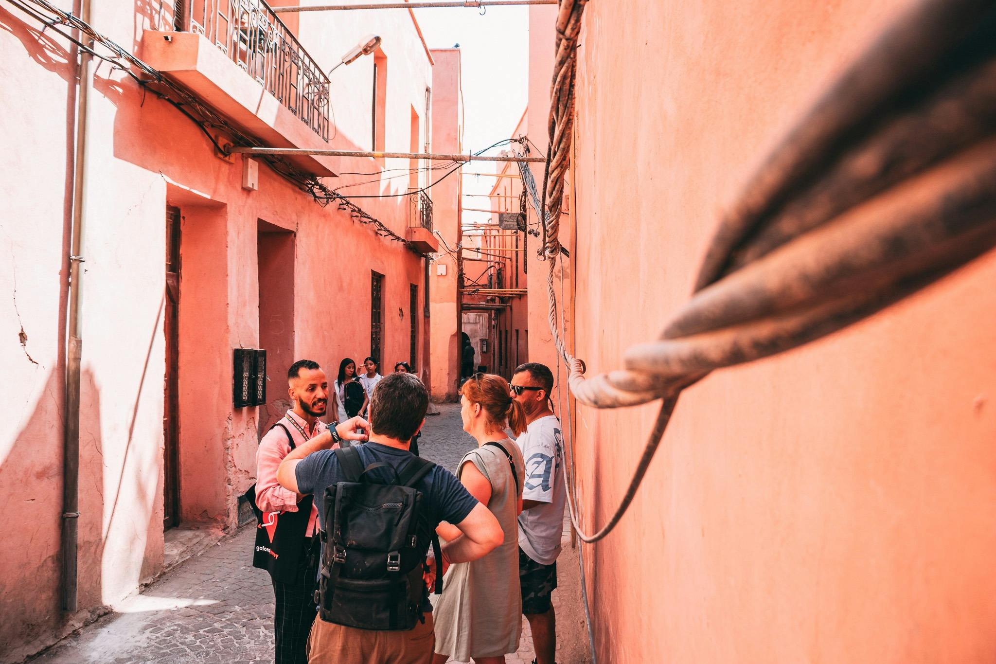 Μια ομάδα τουριστών συνομιλεί σε ένα στενό δρομάκι με ροζ τοίχους, εκτεθειμένα καλώδια και μικρά παράθυρα κάτω από το απογευματινό φως του ήλιου.