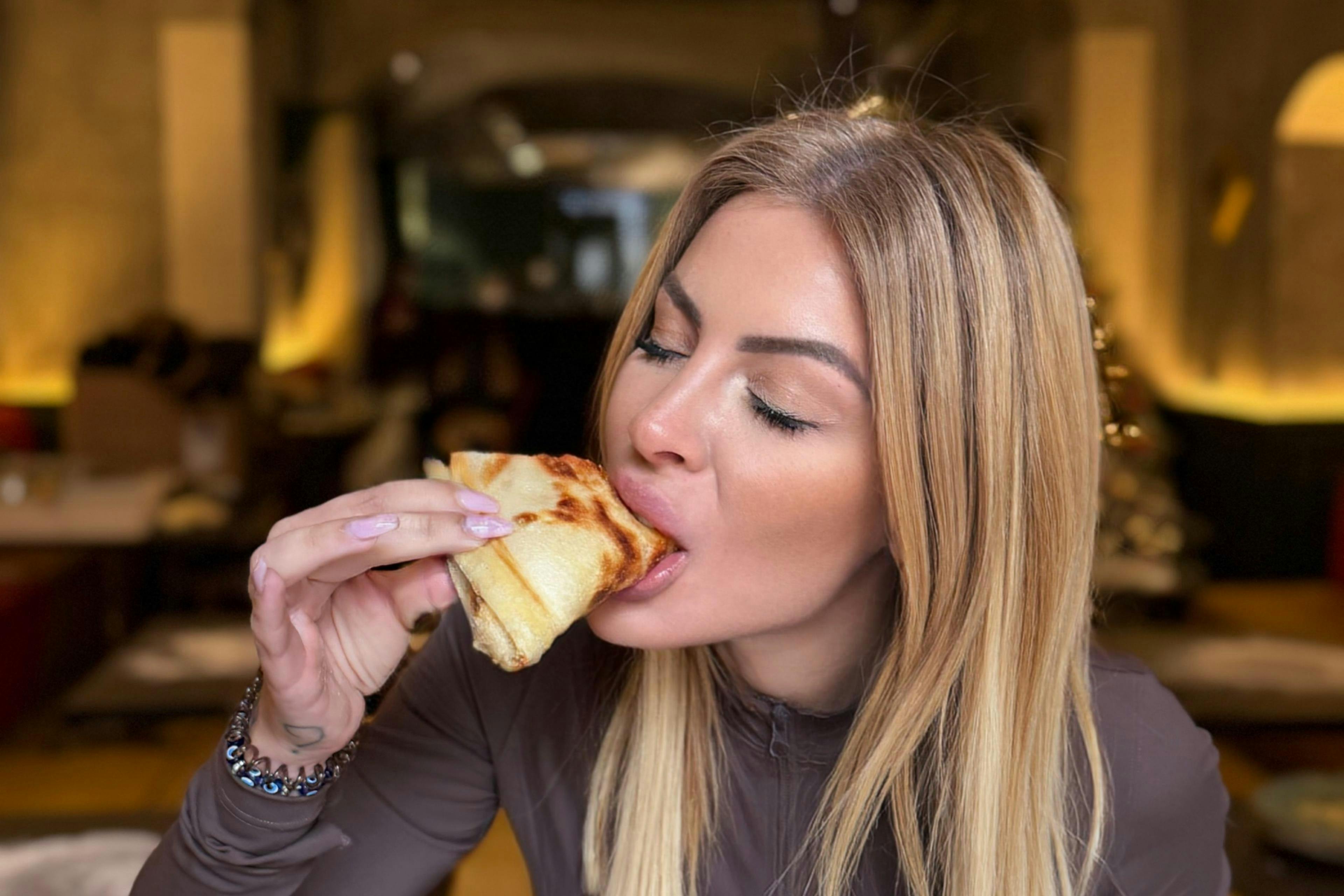 A woman with long blonde hair eating a folded pancake in a dimly lit indoor setting.