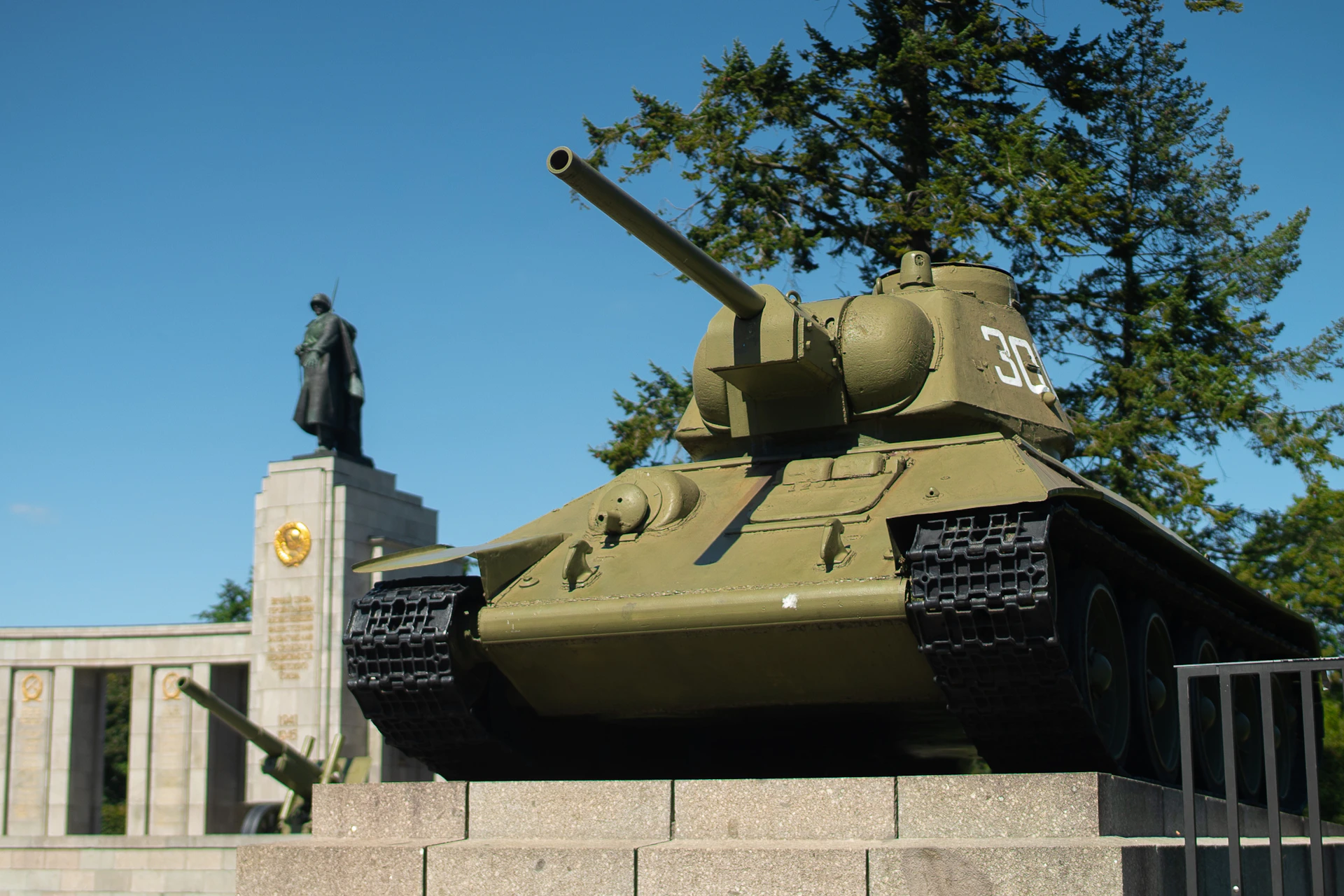 A green military tank on a stone pedestal with a statue of a soldier in the background, under a clear blue sky.