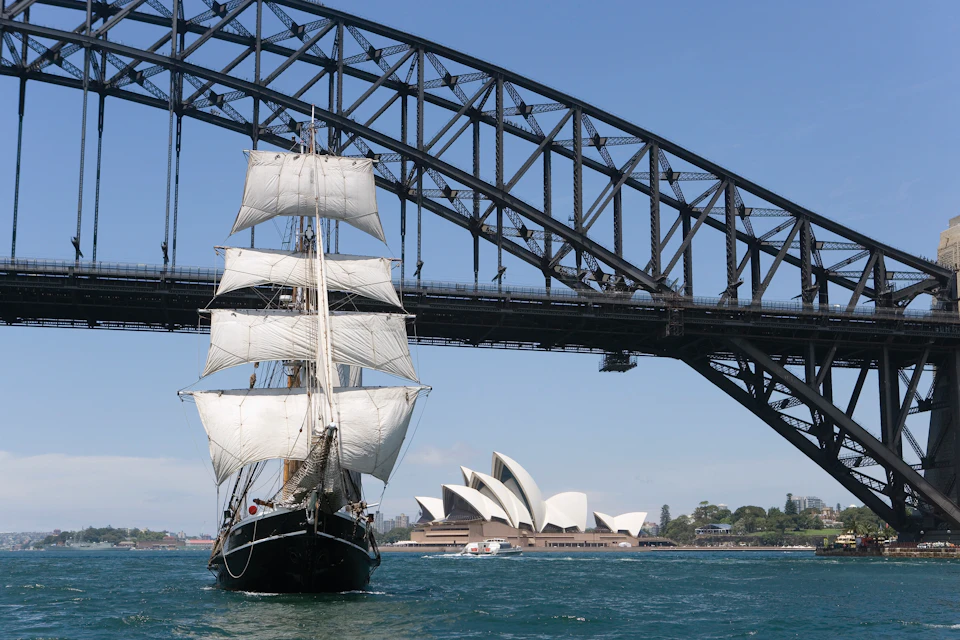 A tall ship with white sails passes under a large steel bridge near a distinctive white building with shell-like structures.