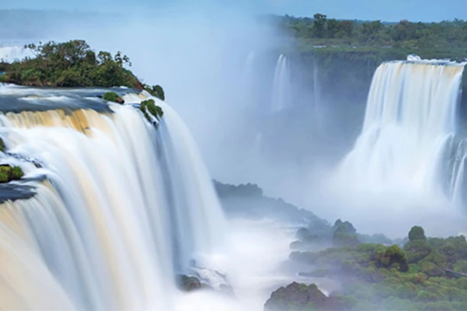 Expansive waterfall cascading over cliffs surrounded by mist, with lush greenery in the background and rocky formations below.