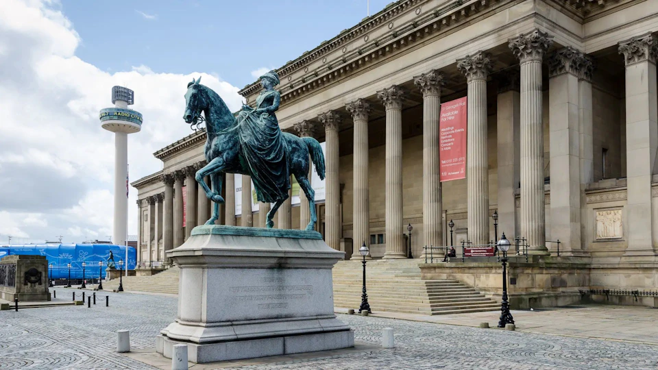 A bronze equestrian statue in front of a grand building with large columns and banners. Cobblestone walkway in the foreground.