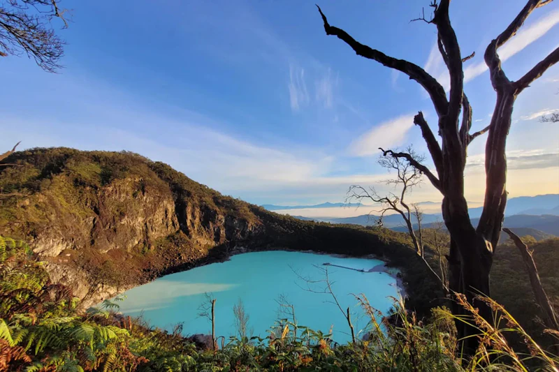 A turquoise lake surrounded by rocky cliffs and vegetation, with a tree silhouette in the foreground under a blue sky.