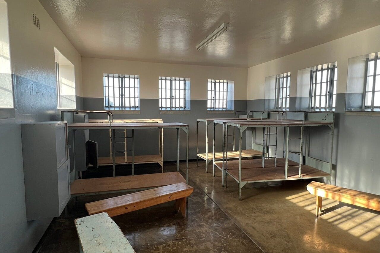 Pokoj s patrovými postelemi s kovovým rámem, dřevěnými lavicemi, zamřížovanými okny a betonovou podlahou.