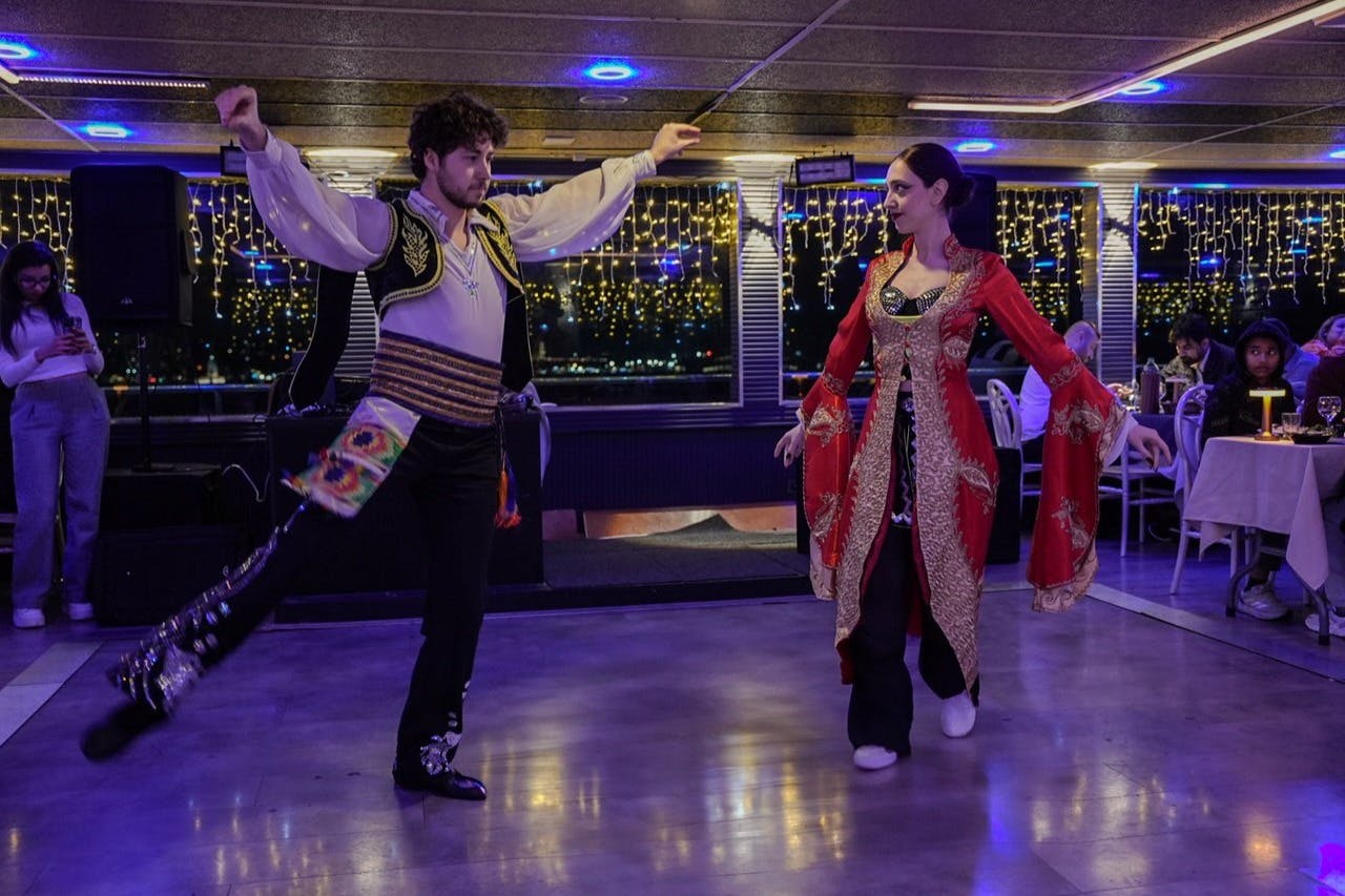 Ein Mann und eine Frau führen einen traditionellen Tanz auf einem gut beleuchteten Boden auf, während im Hintergrund Menschen an Tischen sitzen.