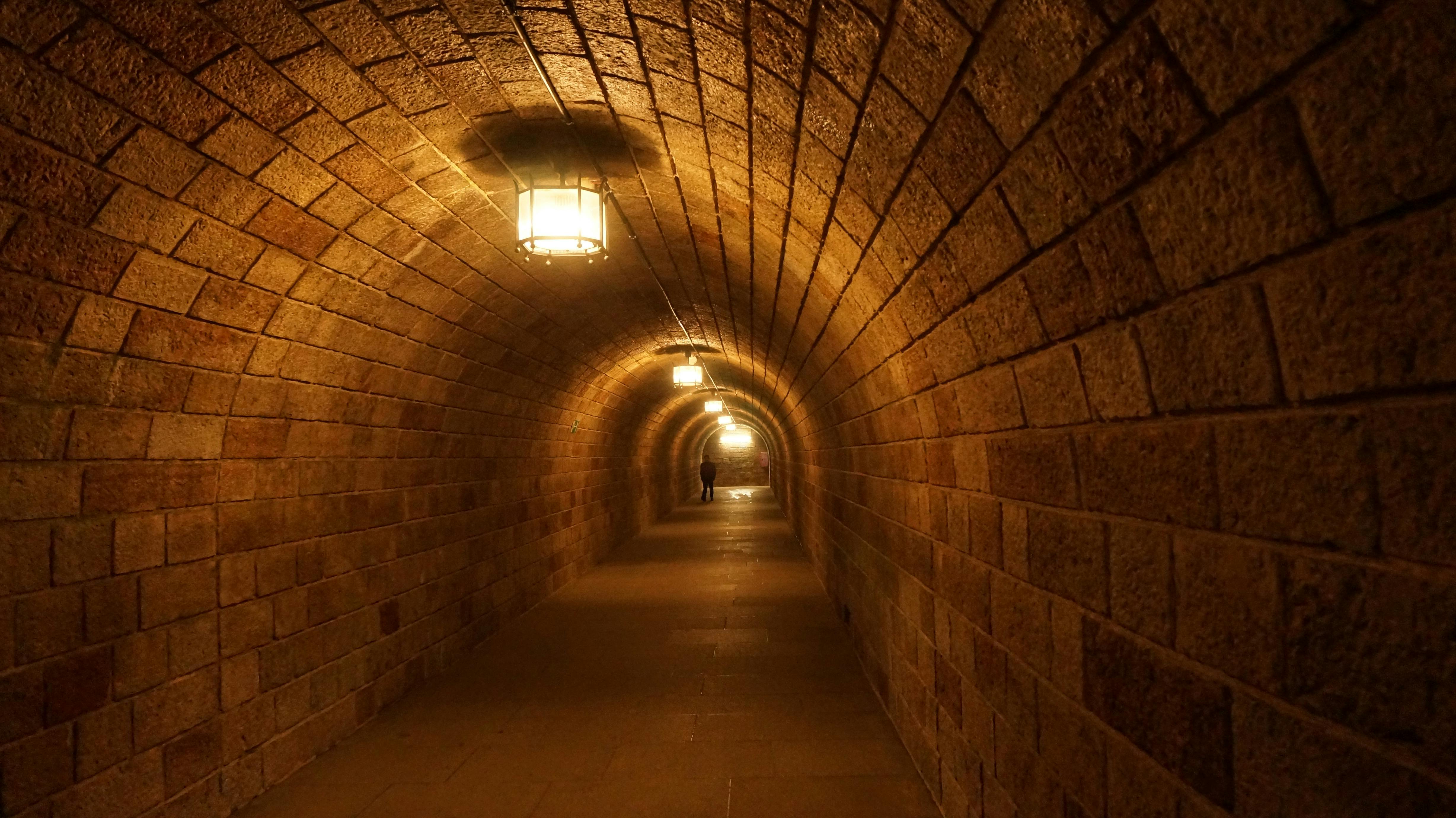 Špatně osvětlený tunel s kamennými stěnami a klenutými stropy, osvětlený závěsnými lucernami. V dálce je vidět osoba.