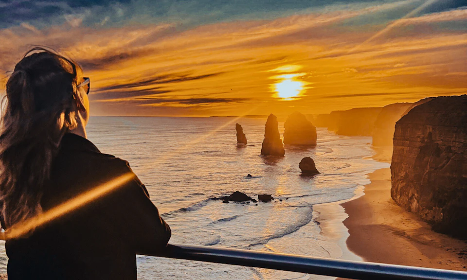 Person leaning on a railing overlooking sea stacks during a vibrant, golden-orange sunset at the ocean.