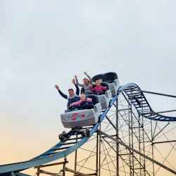 Four people riding a rollercoaster with raised hands and excited expressions as the car descends. Sky and coaster track in the background.