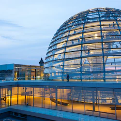 Modern glaskupolstruktur i skymningen med människor som går runt, upplyst interiör och historiska byggnader i bakgrunden.