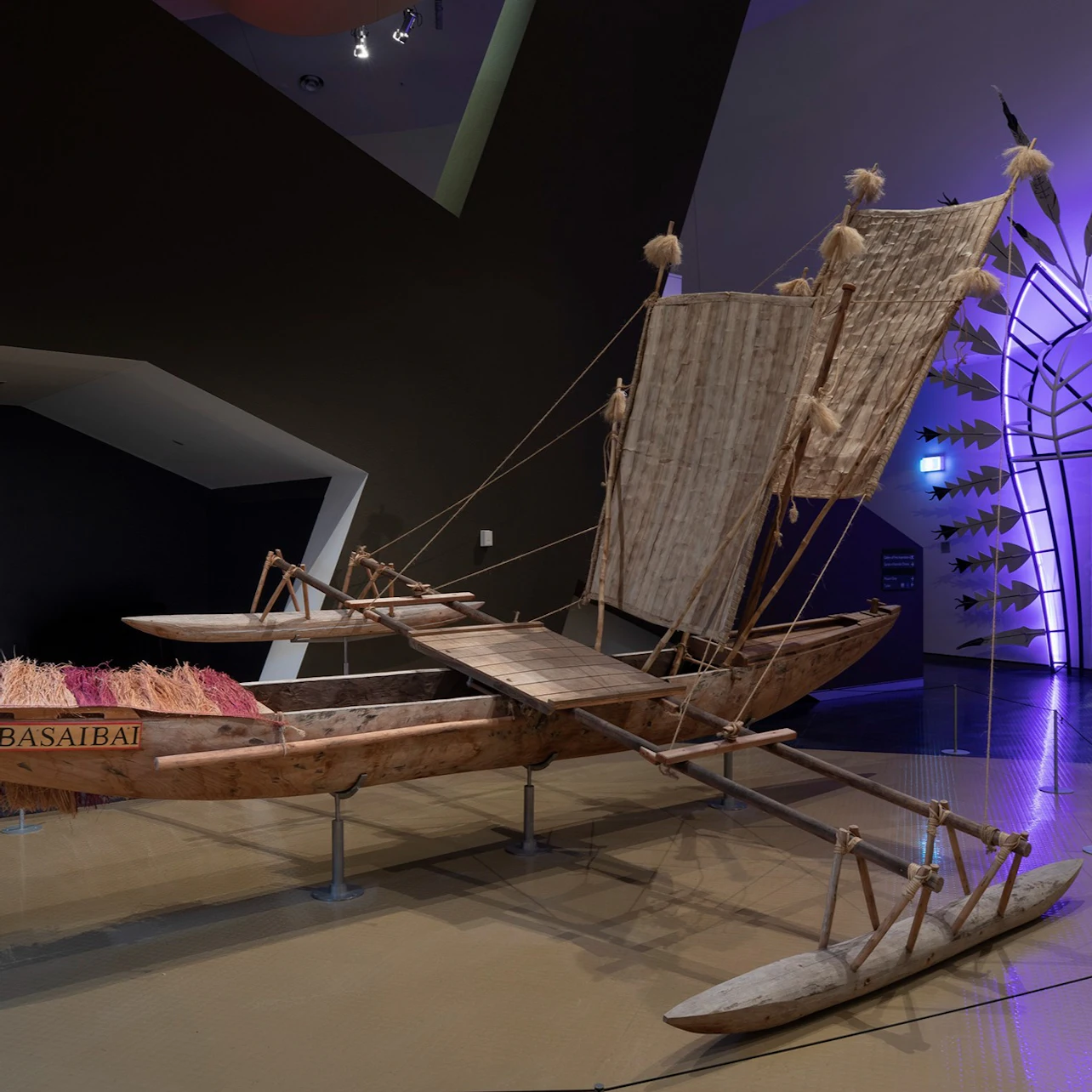 A traditional outrigger canoe with sails displayed in a modern museum exhibit, lit with focused lighting.