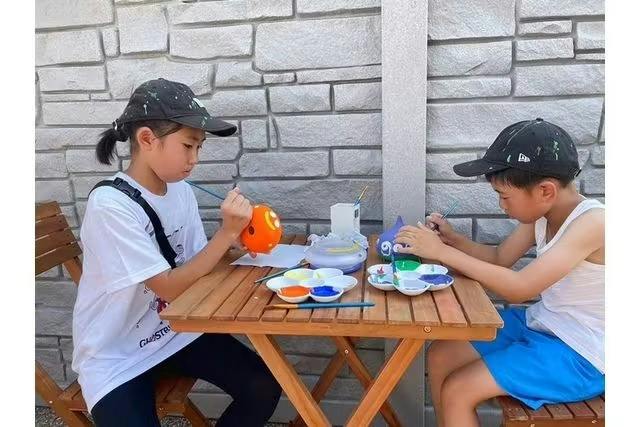 A child painting a small orange object at a wooden table with paint supplies outdoors near a stone wall.