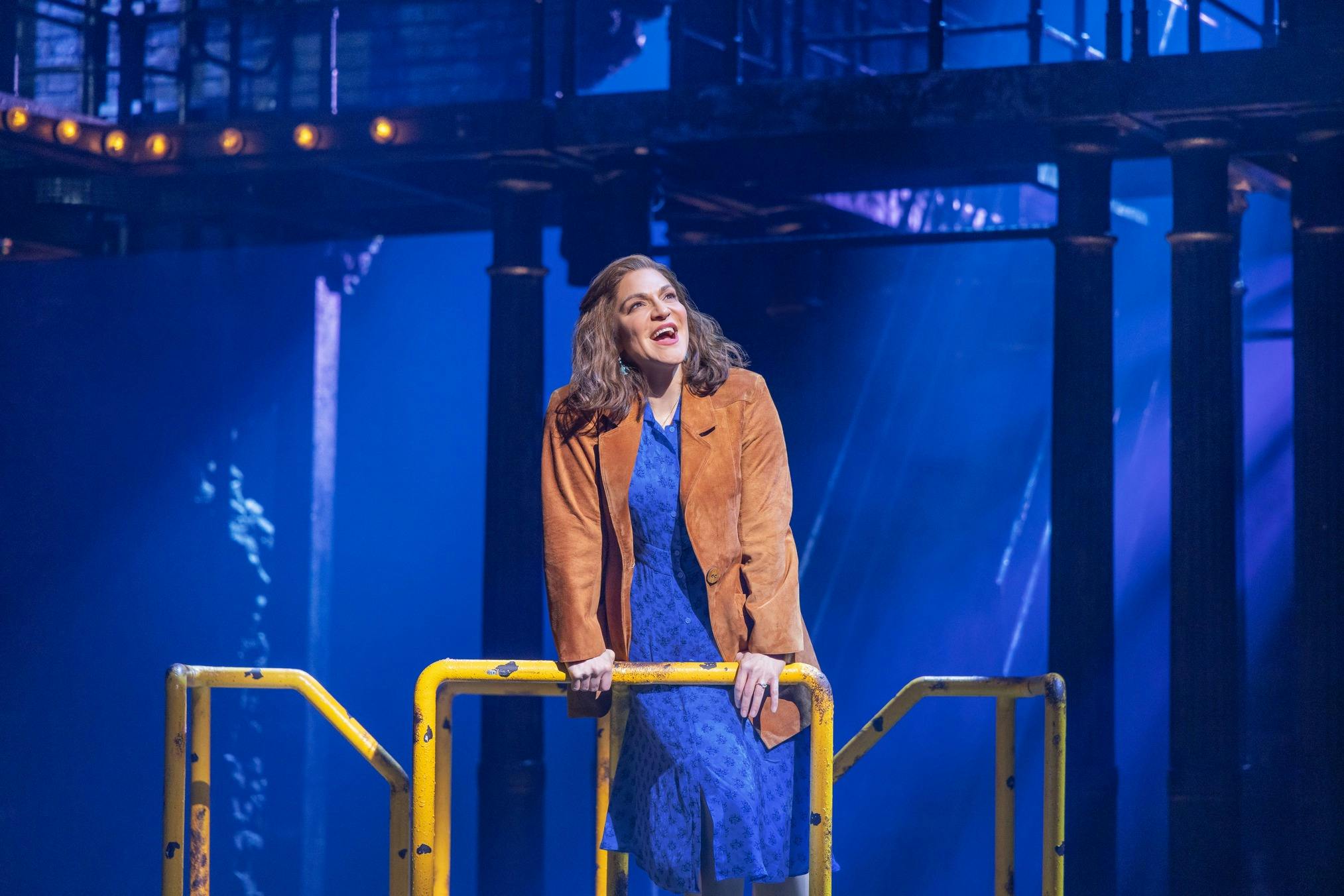 A woman in a blue dress and brown jacket stands behind a yellow railing on a stage with a blue-lit industrial setting.