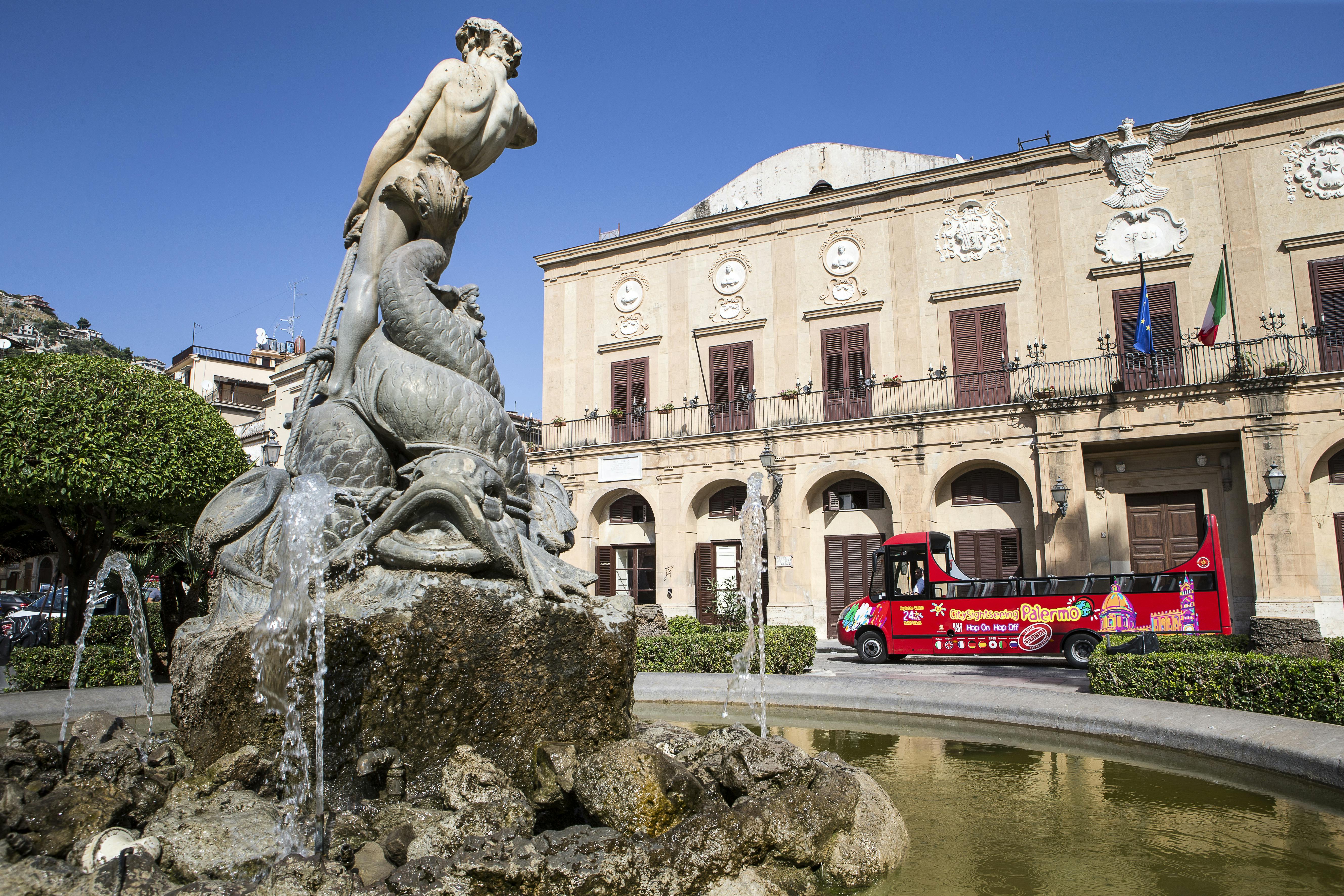 A stone fountain with a statue in the foreground, a colorful tour bus, and a historic beige building under a clear blue sky.