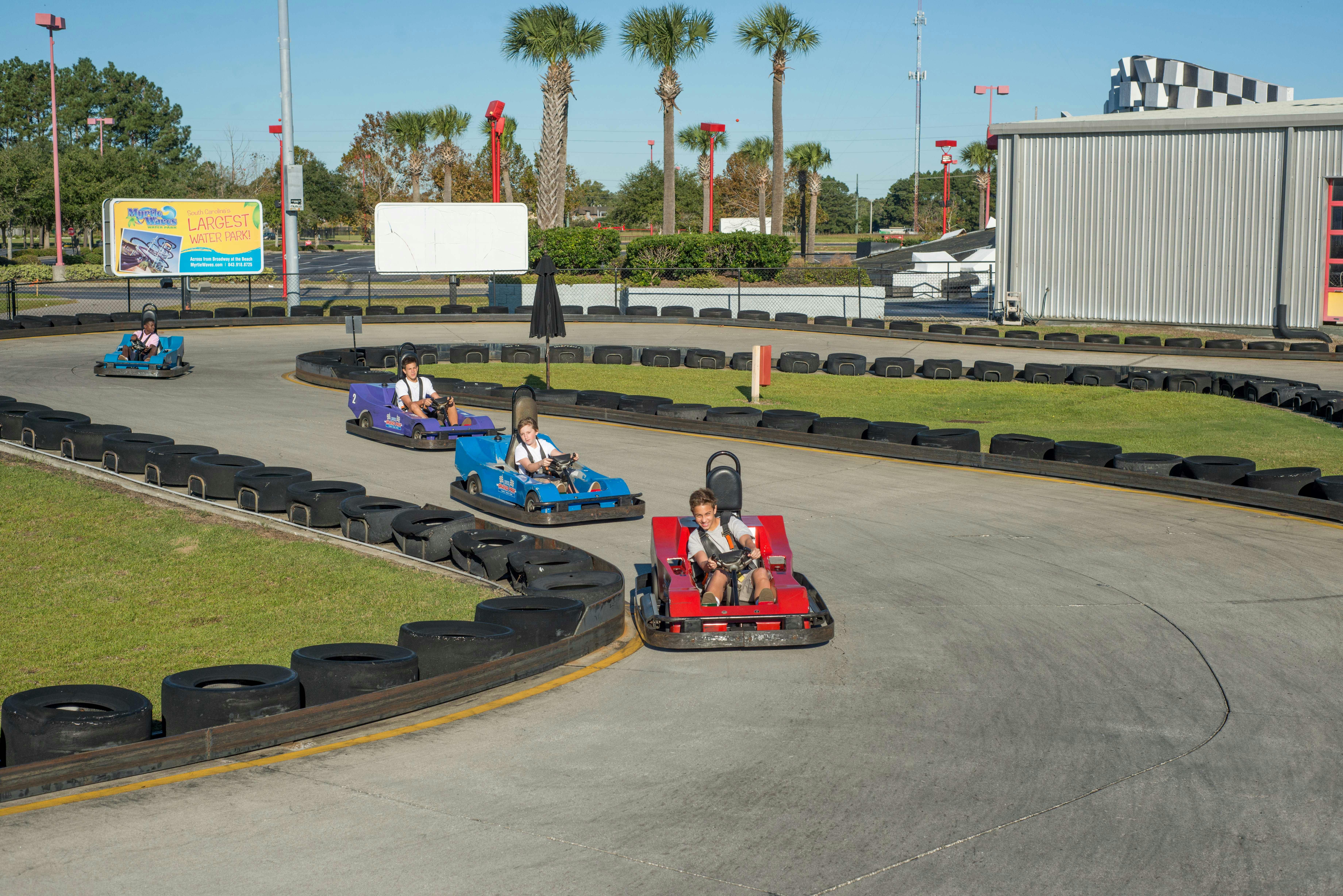 Three people driving red and blue go-karts on a track lined with black tires, with palm trees and buildings in the background.