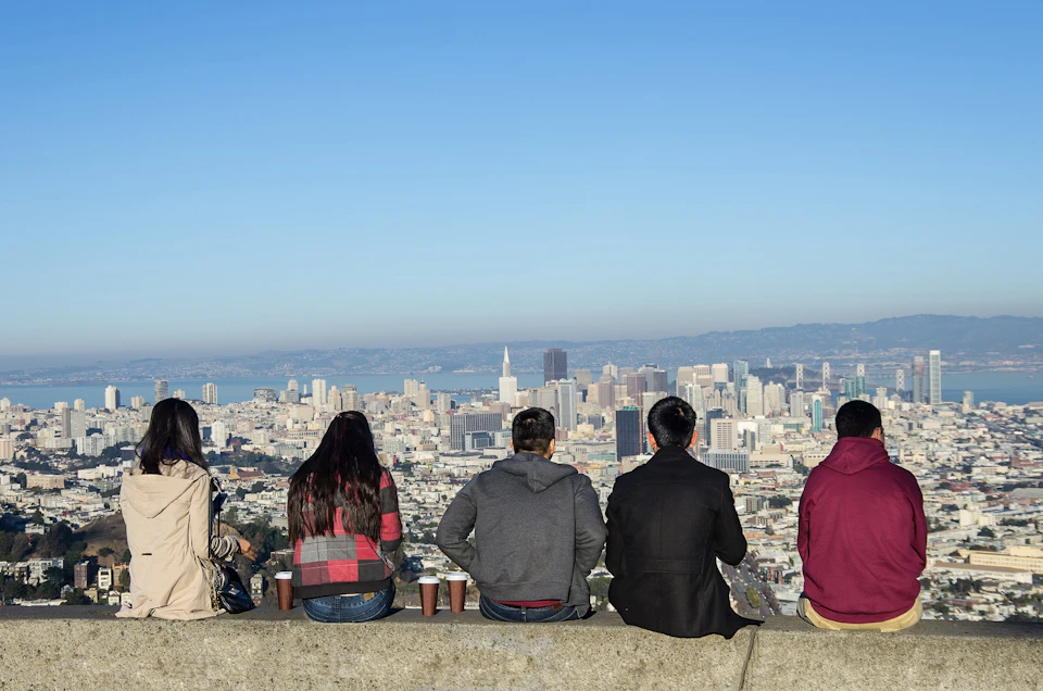 Five people sitting on a ledge overlooking a cityscape with skyscrapers and a clear blue sky in the background.
