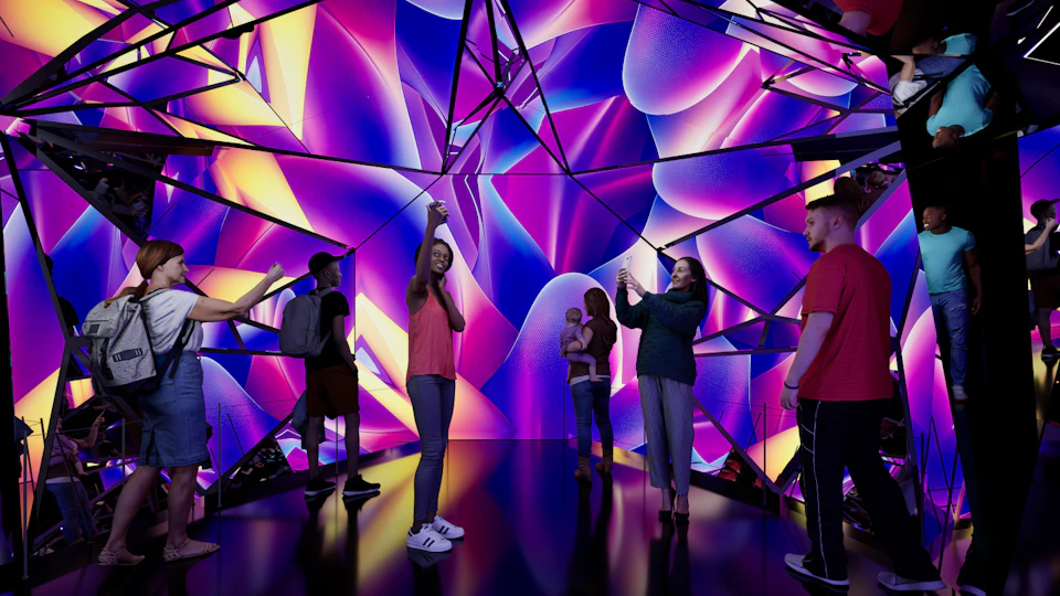 People standing in a vibrant, mirrored room with colorful abstract lighting and reflections, some taking photos.
