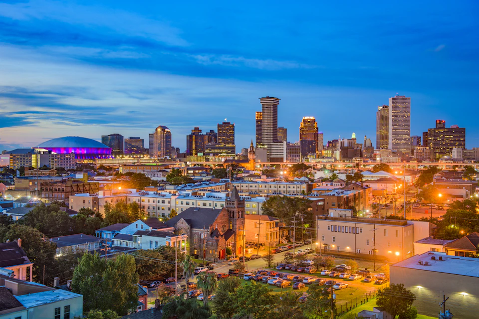 Cityscape at dusk with illuminated buildings, a church, and a parking lot in the foreground, and a stadium in the background.