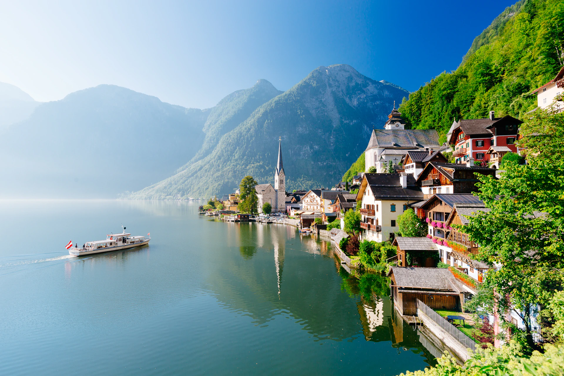 A lakeside village with colorful buildings and a church steeple, set against a backdrop of mountains under a clear blue sky.