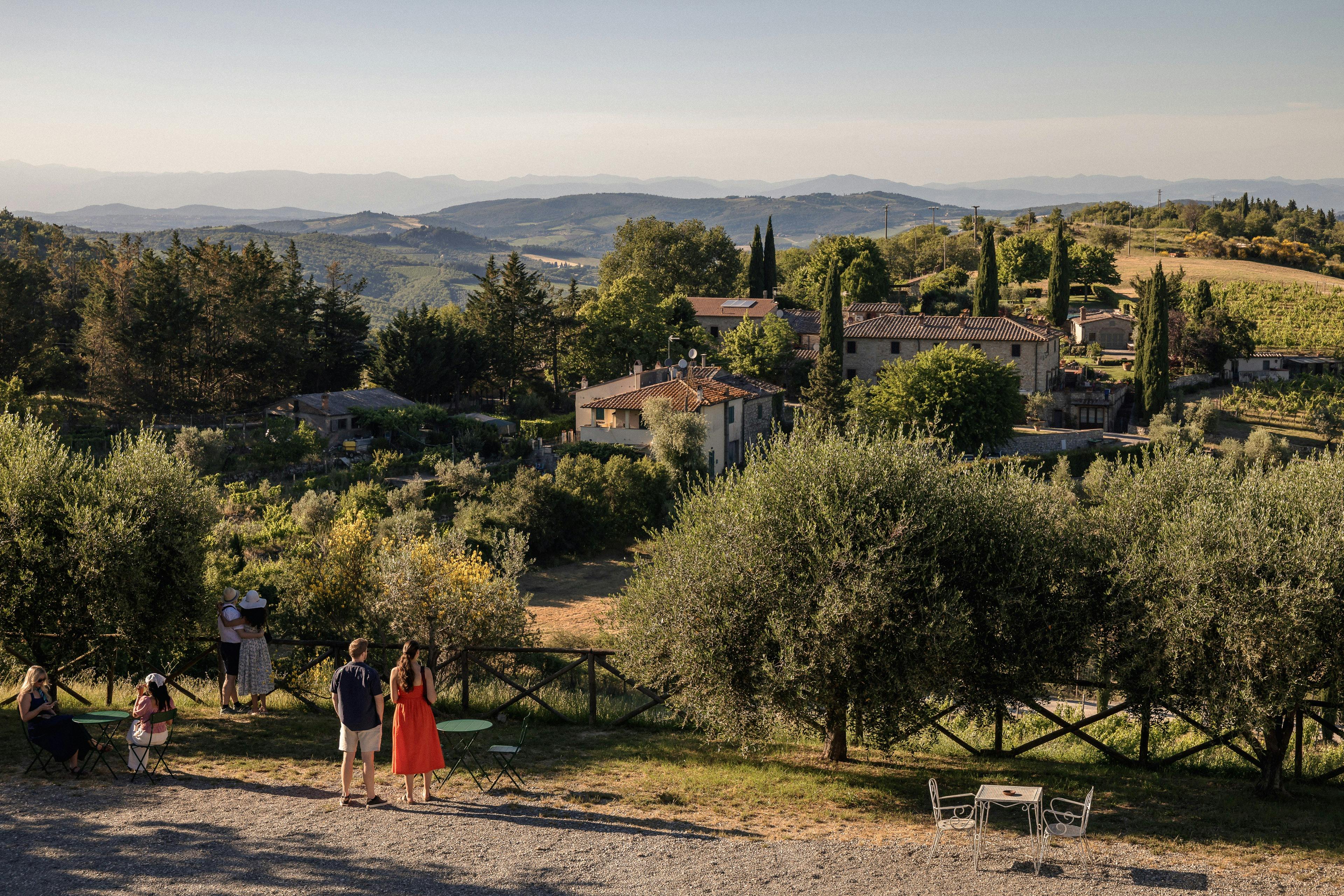 Two people stand near a wooden fence overlooking a rural village with houses, trees, and rolling hills in the background.