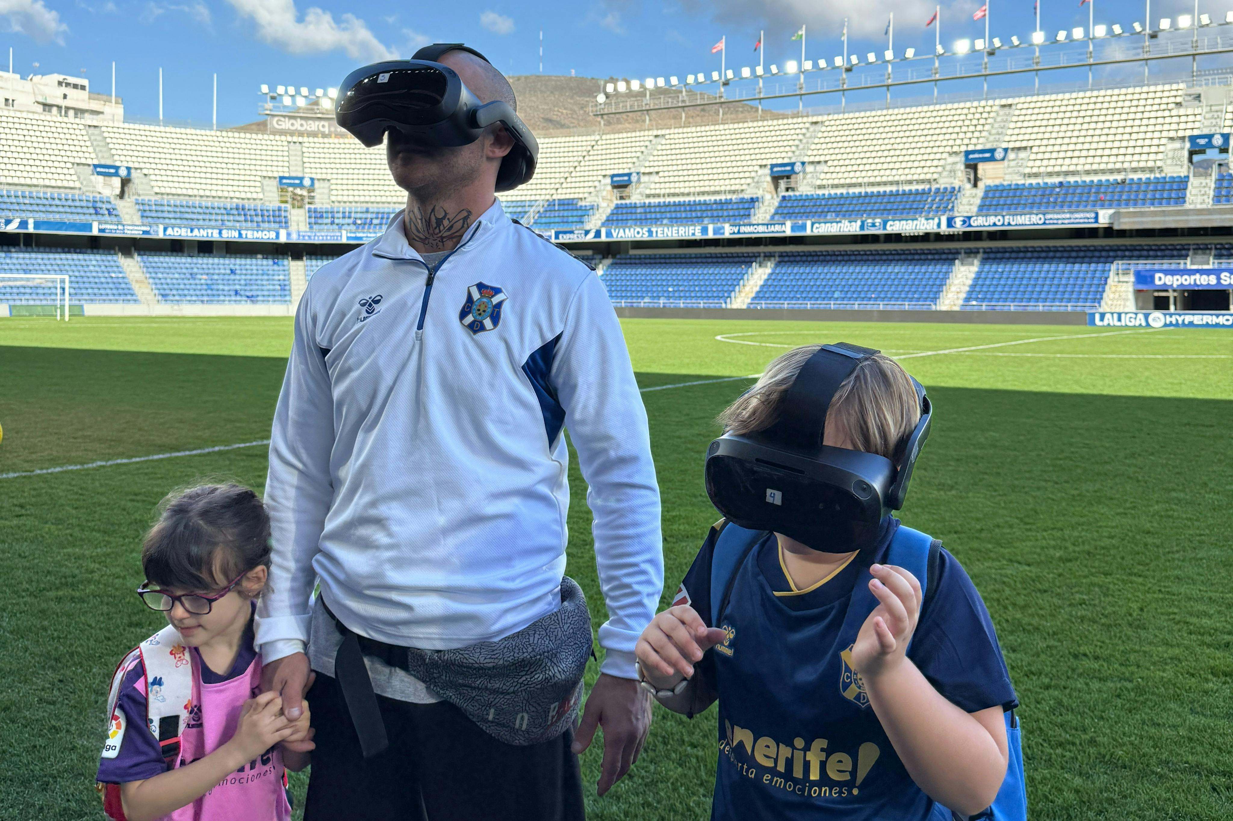 バーチャルリアリティヘッドセットを着用した2人の子供と1人の大人が、スタジアムの座席を背景にしたサッカー場に立っています。