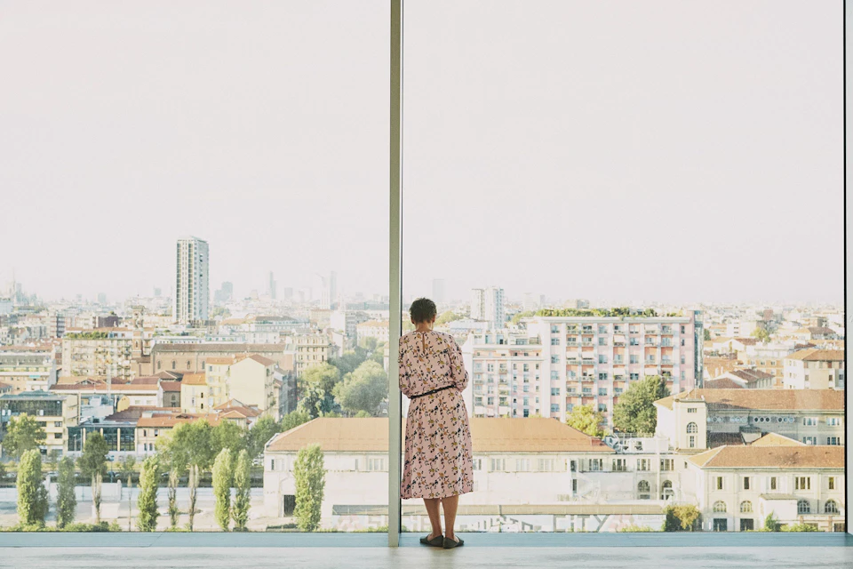 A person in a floral dress is standing by a large window overlooking a cityscape featuring buildings, trees and a clear sky.